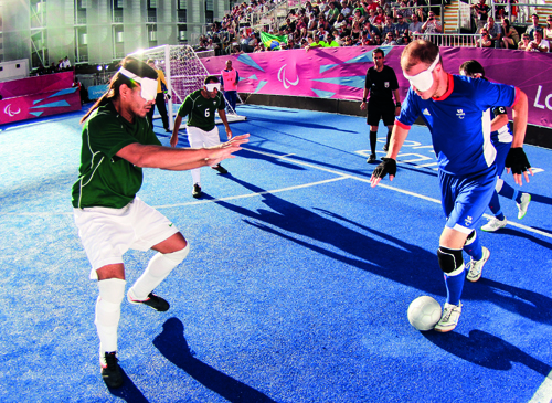 Match de cécifoot lors d’une compétition paralympique, deux joueurs malvoyants s’affrontent balle au pied sur un terrain bleu sous les yeux du public.