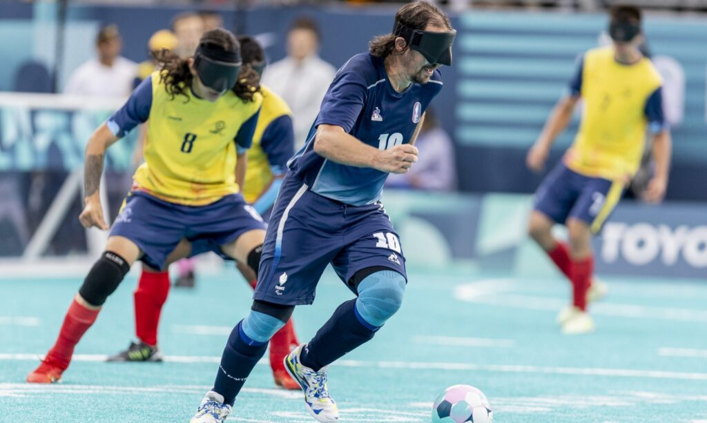 Joueur de l’équipe de France de football pour aveugles en action lors d’un match officiel, portant un bandeau opaque et contrôlant le ballon avec précision sur un terrain indoor.
